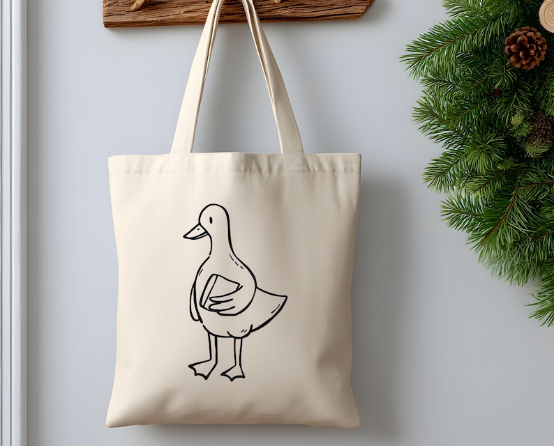 A beige canvas tote bag hanging on a wall next to festive evergreen décor. The bag displays a hand-drawn goose holding a book, printed in simple black line art. The mockup highlights how the bookish goose SVG works beautifully on tote bags, bookstore merchandise, reusable shopping bags, and cozy aesthetic gifts for readers. Clean lines make the design suitable for screen printing, heat transfer vinyl, and sublimation projects.