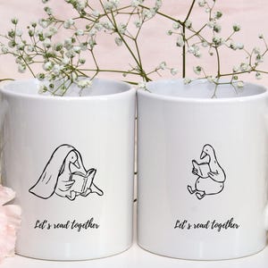Two white ceramic mugs displayed among soft floral decorations. Each mug features a cozy bookish goose illustration—one reading under a blanket and the other sitting and reading calmly. Both mugs include the phrase “Let’s read together,” creating a warm and friendly reading vibe. This mockup demonstrates how the SVG and PNG files can be used for mug designs, book club gifts, café merchandise, or cozy reading-themed products.