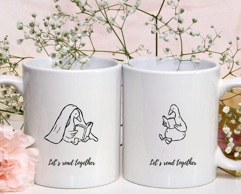 Two white ceramic mugs displayed among soft floral decorations. Each mug features a cozy bookish goose illustration—one reading under a blanket and the other sitting and reading calmly. Both mugs include the phrase “Let’s read together,” creating a warm and friendly reading vibe. This mockup demonstrates how the SVG and PNG files can be used for mug designs, book club gifts, café merchandise, or cozy reading-themed products.
