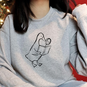 A gray sweatshirt featuring a black line-art illustration of a goose holding a book and magnifying glass from the bookish goose collection. The model sits in a festive cozy setting with Christmas lights in the background. The clean minimal line art stands out clearly on fabric, showing how the SVG can be used for apparel printing, Cricut iron-on designs, book lover merch, and cozy reading-themed clothing for small shops and handmade businesses.