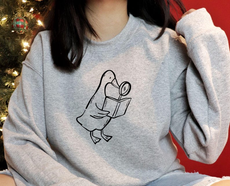 A gray sweatshirt featuring a black line-art illustration of a goose holding a book and magnifying glass from the bookish goose collection. The model sits in a festive cozy setting with Christmas lights in the background. The clean minimal line art stands out clearly on fabric, showing how the SVG can be used for apparel printing, Cricut iron-on designs, book lover merch, and cozy reading-themed clothing for small shops and handmade businesses.