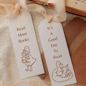 Two printed bookmarks lying on a soft beige fabric, each featuring hand-drawn reading goose illustrations. One bookmark shows a goose carrying a tall stack of books with the quote “Read More Books,” while the other shows a goose reading while sitting on a cushion with the quote “It’s A Good Day To Read.” The warm tones and textured paper create a cozy atmosphere, showcasing how the SVG and PNG files can be used for bookish stationery, gifts, and printable crafts.