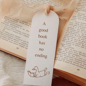 A bookmark mockup featuring a hand-drawn goose reading under a blanket printed in warm brown line art on textured ivory paper. The bookmark includes the quote “A good book has no ending” and is tied with a soft beige ribbon. The scene is placed on an open vintage book, creating a cozy reading aesthetic. This image demonstrates how the bookish goose SVG and PNG files can be used for printed bookmarks, bookish gifts, book club merch, and handmade stationery.