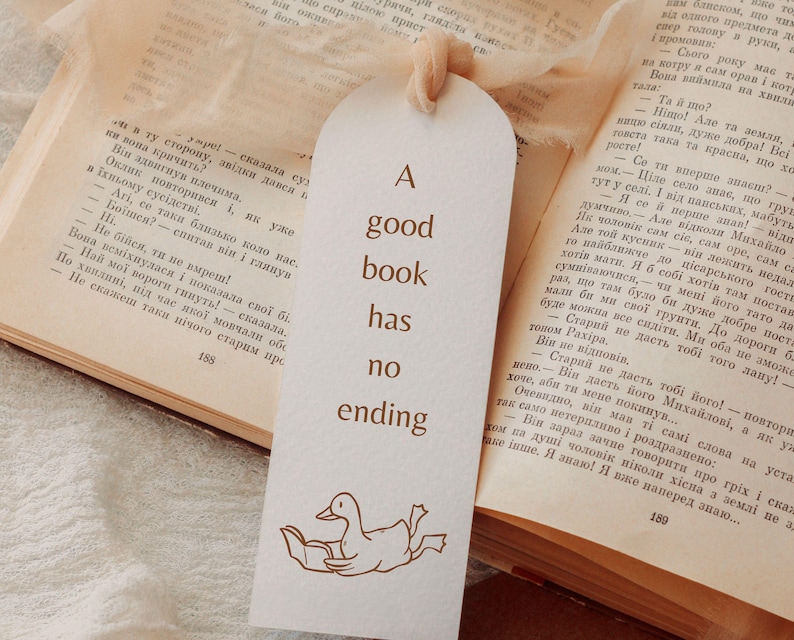 A bookmark mockup featuring a hand-drawn goose reading under a blanket printed in warm brown line art on textured ivory paper. The bookmark includes the quote “A good book has no ending” and is tied with a soft beige ribbon. The scene is placed on an open vintage book, creating a cozy reading aesthetic. This image demonstrates how the bookish goose SVG and PNG files can be used for printed bookmarks, bookish gifts, book club merch, and handmade stationery.