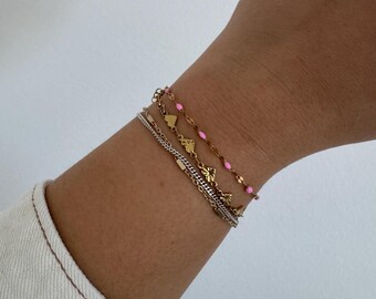Könnte beinhalten: Ein Set aus vier goldenen Armbändern mit rosa Perlen und kleinen goldenen Anhängern. Die Armbänder sind übereinander am Handgelenk einer Person gelegt.