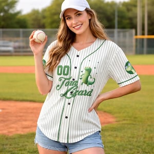 Puede incluir: Camiseta de béisbol blanca con rayas verdes, con el texto "Toid Lizard" y un dibujo de un lagarto de dibujos animados. La camiseta tiene una parte delantera abotonada y detalles verdes. También se ve una gorra de béisbol blanca y una pelota de béisbol.