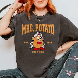 Puede incluir: Una camiseta gris oscuro con el texto "MRS. POTATO" en letras amarillas y naranjas. La camiseta también presenta una imagen de dibujos animados de la Sra. Potato Head de la franquicia Toy Story, con el texto "EST. 1999 TOY STORY" debajo de la imagen.