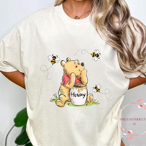 May include: A white t-shirt with a cartoon image of Winnie the Pooh holding a jar of honey that says "Hunny". There are three cartoon bees flying around Winnie the Pooh.