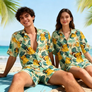 Puede incluir: Conjunto a juego de camisa y pantalón corto con estampado tropical. El estampado presenta ilustraciones de cachorros de león amarillos, hojas verdes y flores naranjas sobre un fondo crema. El conjunto lo llevan dos personas en una playa.