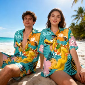 Puede incluir: Conjunto a juego de camisas y pantalones cortos hawaianos turquesa, naranja y rosa con Pluto. Las camisas y los pantalones cortos tienen un diseño floral tropical con gráficos de Pluto y la palabra "Pluto" impresa en la camisa. La pareja está sentada en una playa de arena.
