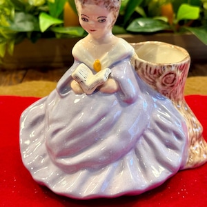 May include: A ceramic figurine of a woman in a lavender dress and hat, holding a book. The figurine has a planter on the right side, designed to look like a tree trunk. The woman's dress is detailed with folds and the book has visible pages.