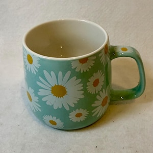May include: A white ceramic mug with a black handle and the text "But First, Coffee" in a bold, black font. The mug is set against a blurred background, suggesting a cozy setting. The mug appears to be approximately 4 inches tall.