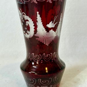 May include: A ruby red glass vase with white etched designs. The vase features a flared top and a bulbous base. The etched designs include swirling patterns and a bird motif, adding a touch of elegance to the decorative piece.