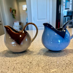 May include: Two ceramic creamer pitchers with a unique glaze design. One pitcher is light gray with brown accents and the other is light blue with dark blue accents.