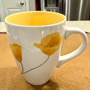 May include: White ceramic mug with a yellow interior and a large, curved handle. The mug is decorated with yellow poppy flower illustrations and green stems. The mug is sitting on a brown surface.