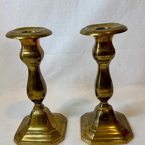 May include: A pair of polished brass candlesticks featuring a wide, circular top rim, a slender, curved central stem, and a square, stepped base. The metal surface exhibits a warm, golden-yellow tone with subtle variations in the reflective finish.