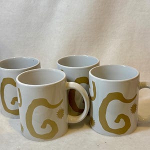 May include: A set of four white ceramic mugs, each featuring a repeating mustard-yellow abstract swirl pattern and small star-like accents. The mugs have a cylindrical shape with a side handle and a smooth, glossy finish on the exterior surface.