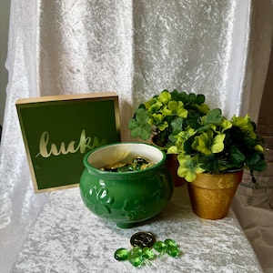 May include: A St. Patrick's Day themed display featuring a green ceramic pot filled with gold coins and green glass beads. A wooden sign with the word "lucky" is next to the pot. Two small gold pots with green and yellow shamrocks are also present.