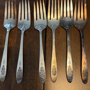 May include: A set of six silver-toned metal forks arranged in a row. Each fork features four tines and a slender handle decorated with a small, intricate engraved pattern near the base. The metal shows varying degrees of tarnish and patina.