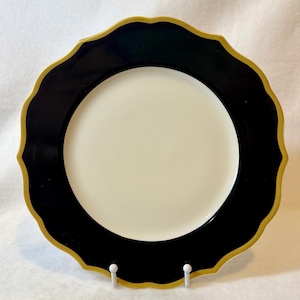 May include: A decorative plate with a scalloped edge. The plate features a black border with a gold trim and a cream-colored center. The plate is displayed on a white stand.