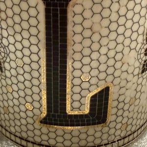May include: Close-up of a cylindrical object with a white honeycomb pattern and a large black letter "L". The letter is outlined in gold and filled with a mosaic-like pattern. Small gold accents are scattered across the surface.