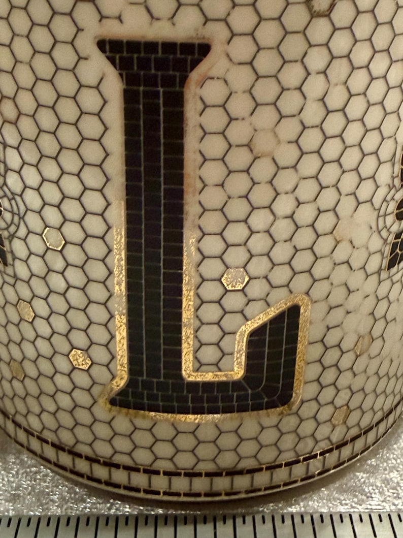 May include: Close-up of a cylindrical object with a white honeycomb pattern and a large black letter "L". The letter is outlined in gold and filled with a mosaic-like pattern. Small gold accents are scattered across the surface.