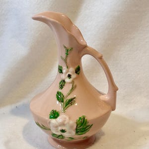 May include: A light pink ceramic pitcher featuring a raised floral design with white flowers and green leaves. The pitcher has a curved handle, a wide base, and a flared spout. The surface is smooth and glossy, showcasing a delicate botanical motif.