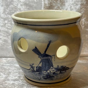 May include: A ceramic jar with a Dutch windmill design. The jar is cream-colored with a blue and white illustration of a windmill and landscape. It has two circular openings and a dark blue rim.
