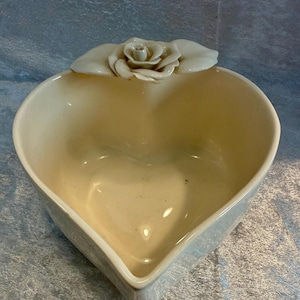 May include: Cream-colored heart-shaped ceramic dish with a rose and leaf accent. The dish has a smooth, glossy finish and a shallow interior. The rose is a light cream color, and the leaves are a similar shade.