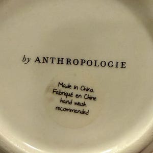 May include: Off-white ceramic item with the text "by ANTHROPOLOGIE" in black. A circular sticker reads "Made in China, Fabriqu&eacute; en Chine, hand wash recommended."