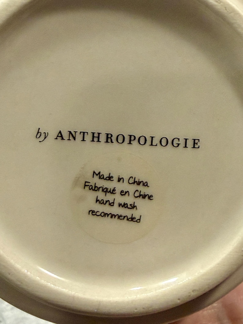 May include: Off-white ceramic item with the text "by ANTHROPOLOGIE" in black. A circular sticker reads "Made in China, Fabriqu&eacute; en Chine, hand wash recommended."