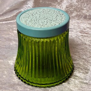 May include: A vintage green glass jar with a ribbed design. The jar has a light blue lid with a floral pattern. The jar is likely used for storage or decoration.