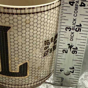May include: A ceramic mug with a mosaic design featuring black and gold accents. The mug has a honeycomb pattern and a floral design. The ruler next to the mug measures approximately 4 inches.