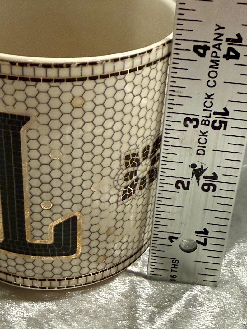 May include: A ceramic mug with a mosaic design featuring black and gold accents. The mug has a honeycomb pattern and a floral design. The ruler next to the mug measures approximately 4 inches.