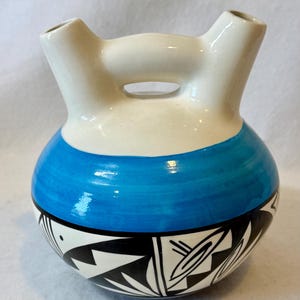 May include: A ceramic, two-spouted vessel with a white top and a blue band around the middle. The lower portion features a black and white geometric design. The vessel has a handle-like opening between the spouts.