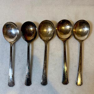 May include: Five vintage silver spoons arranged in a row. The spoons have oval bowls and long, slender handles with decorative detailing near the base. The spoons show signs of age and use, with some tarnishing visible on the silver surface.