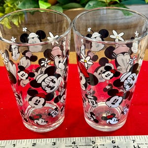 Puede incluir: Dos vasos transparentes decorados con un patr&oacute;n repetido de Minnie Mouse y dise&ntilde;os florales en negro, blanco y rojo. Los vasos son cil&iacute;ndricos y est&aacute;n sobre una superficie roja.