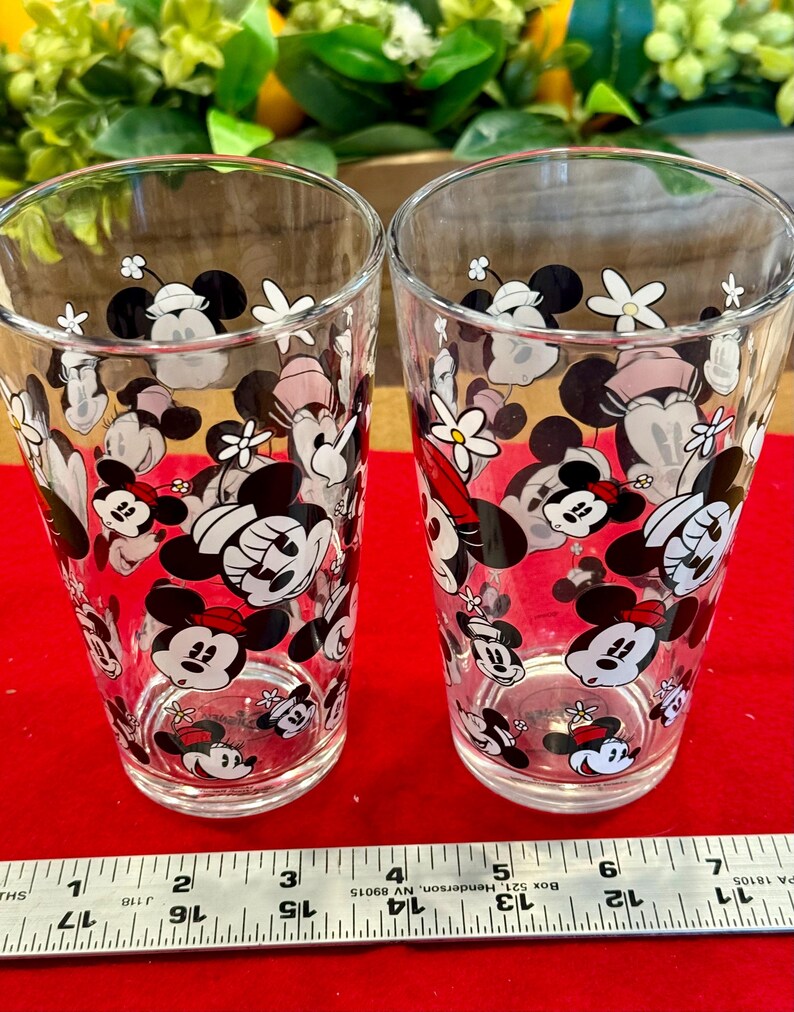 Puede incluir: Dos vasos transparentes decorados con un patr&oacute;n repetido de Minnie Mouse y dise&ntilde;os florales en negro, blanco y rojo. Los vasos son cil&iacute;ndricos y est&aacute;n sobre una superficie roja.