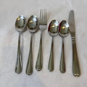 May include: A set of stainless steel silverware including spoons, a fork, and a knife. The spoons have a teardrop-shaped bowl and a long, slender handle. The fork has four tines, and the knife has a rounded blade. The silverware is arranged on a white surface.