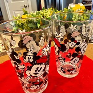 Puede incluir: Dos vasos transparentes con un patr&oacute;n repetido de caras de Mickey Mouse y Minnie Mouse, flores y lunares. Los vasos est&aacute;n sobre una superficie roja.