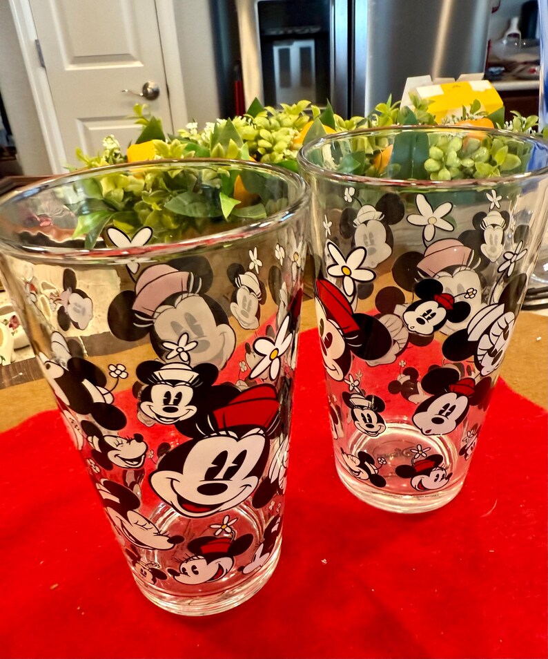 Puede incluir: Dos vasos transparentes con un patr&oacute;n repetido de caras de Mickey Mouse y Minnie Mouse, flores y lunares. Los vasos est&aacute;n sobre una superficie roja.