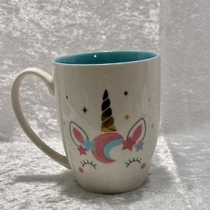 May include: White ceramic mug with a teal interior and a unicorn face design. The unicorn features a black and gold horn, pink and blue accents, and star details. The mug has a curved handle.