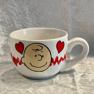 May include: White ceramic mug featuring a cartoon character's face with a smiling expression, red hearts, and a red zigzag design. The mug has a curved handle and is suitable for hot or cold beverages.