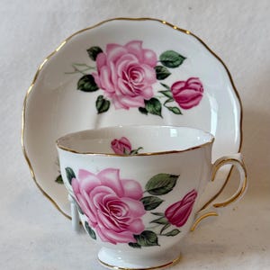 May include: A white teacup and saucer set featuring pink rose floral designs and gold trim. The teacup has a pedestal base and a curved handle. The saucer has a scalloped edge. The set is designed for tea parties.