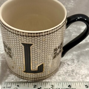 May include: A ceramic mug with a black handle and a cream-colored interior. The exterior features a honeycomb pattern with the letter "L" in black and gold. The mug has a black rim and gold accents.