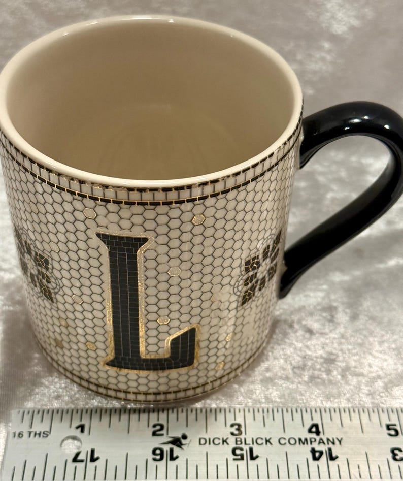 May include: A ceramic mug with a black handle and a cream-colored interior. The exterior features a honeycomb pattern with the letter "L" in black and gold. The mug has a black rim and gold accents.