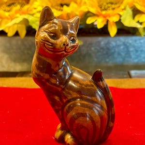 May include: A ceramic cat figurine in shades of brown with darker stripes. The cat is posed sitting upright with a smiling expression. The figurine is on a red surface, with yellow flowers in the background.