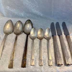 May include: A collection of vintage silverware, including spoons and knives, arranged on a light blue surface. The spoons vary in size, and the knives have serrated blades. The silverware has decorative detailing on the handles.