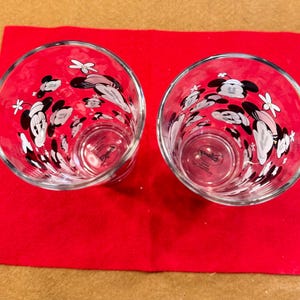 Puede incluir: Dos vasos transparentes con un dise&ntilde;o de Mickey Mouse, con detalles en negro, blanco y rosa. Los vasos est&aacute;n colocados sobre una superficie roja. El dise&ntilde;o incluye la cara de Mickey Mouse y otros elementos decorativos.