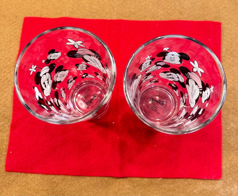 Puede incluir: Dos vasos transparentes con un dise&ntilde;o de Mickey Mouse, con detalles en negro, blanco y rosa. Los vasos est&aacute;n colocados sobre una superficie roja. El dise&ntilde;o incluye la cara de Mickey Mouse y otros elementos decorativos.